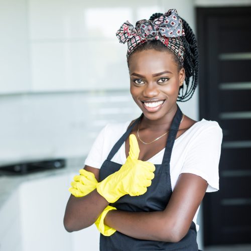 household, cleaning and people concept happy afro amerocanwoman or housewife in rubber gloves wiping table with microfiber cloth at home kitchen and showing thumbs up
