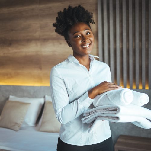 african american house keeper at a guest bedroom holding towels while facing camera smiling. cleaning the hotel room. hotel vacations. smiling housekeeping woman
